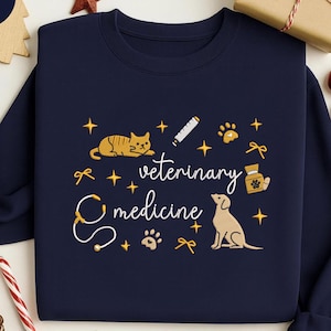 Könnte beinhalten: Marineblaues Sweatshirt mit einem Tiermedizin-Design. Das Design umfasst eine Katze, einen Hund, ein Stethoskop, Pfotenabdrücke und die Worte "Tiermedizin". Goldene Sterne und Schleifen vervollständigen das Design.