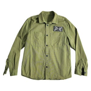 May include: An olive green long-sleeve button-down shirt featuring a pointed collar, two chest pockets, and a rectangular black and white patch on the upper left chest that displays the text HURRICANE inside a stylized Union Jack flag design.
