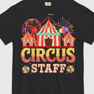 以下が含まれることがあります： 黒いTシャツ。カラフルなサーカスのデザインが特徴です。観覧車、サーカスのテント、風船、そして「CIRCUS STAFF」の文字が装飾的なフォントで描かれています。デザインは赤、黄、青、緑の色合いを使用しています。