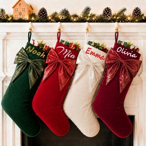 May include: Four velvet Christmas stockings hanging in a row. The first is dark green with the name Noah, the second is red with the name Mia, the third is white with the name Emma, and the fourth is burgundy with the name Olivia. Each has a large bow.