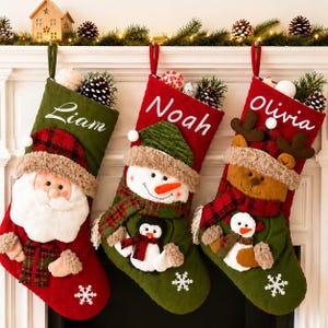 May include: Three Christmas stockings hanging in a row. The first is green with the name Liam and a Santa Claus applique. The second is red with the name Noah and a snowman applique. The third is red with the name Olivia and a reindeer applique.