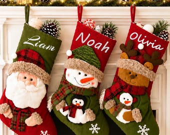 Op de afbeelding: Drie kerstsokken hangen op een rij. De eerste is groen met de naam Liam en een kerstman-applicatie. De tweede is rood met de naam Noah en een sneeuwpop-applicatie. De derde is rood met de naam Olivia en een rendier-applicatie.
