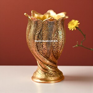 Könnte beinhalten: Verzierte goldfarbene Vase mit strukturierter Oberfläche und aufwendigen floralen Mustern. Die Vase hat eine ausgestellte Oberseite und einen Sockel. Der Text "Mystic Handicraft Arts" ist sichtbar. Rechts von der Vase befindet sich eine gelbe Blume.