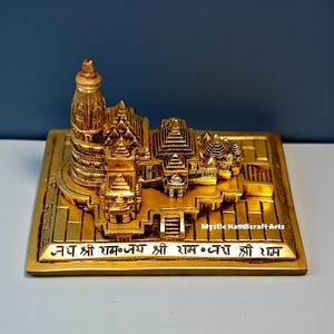 May include: A gold-coloured, detailed miniature temple sculpture. The intricate design features multiple towers, domes, and architectural elements, set on a rectangular base with engraved text. The base also has the text "Mystic Handicraft Arts".
