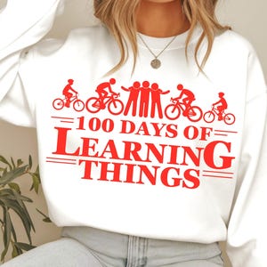 May include: White sweatshirt with red graphic text and illustrations. The text reads "100 DAYS OF LEARNING THINGS" with bicycle and people illustrations. A casual garment.