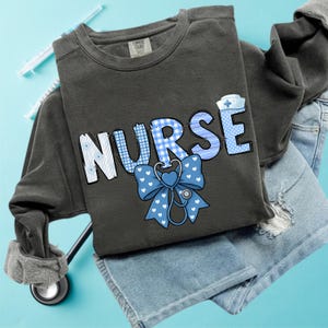 May include: A dark grey sweatshirt featuring the word "NURSE" in blue and white lettering. The letter "U" has a blue and white gingham pattern. A blue bow with white hearts and a stethoscope graphic are below the word. The sweatshirt is folded on blue jeans.