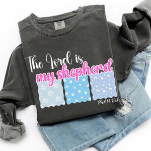 Może przedstawiać: Ciemnoszara bluza z napisem "The Lord is my shepherd" w kolorze białym i różowym. Poniżej znajdują się trzy prostokąty z falistymi brzegami z wzorami stokrotek w kolorze jasnoniebieskim i niebieskim. Tekst "Psalm 23:1" znajduje się poniżej.