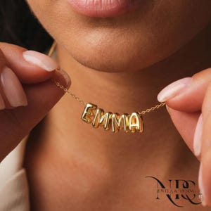 Könnte beinhalten: Eine goldfarbene Halskette mit dem Namen "EMMA" in Großbuchstaben. Die Kette wird von einer Person gehalten, die die zarte Kette und die einzelnen Buchstabenperlen zeigt. Der Schmuck ist von der Marke "NID JEWELS & DESING".