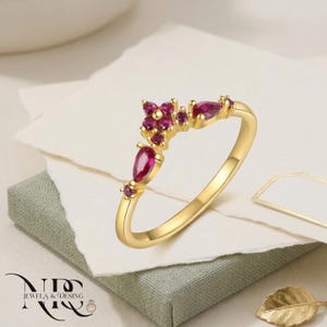 May include: A gold ring with a floral design, set with small, round and teardrop-shaped red gemstones. The ring is displayed on a white surface with a gold leaf and the text "NID JEWELS & DESING".