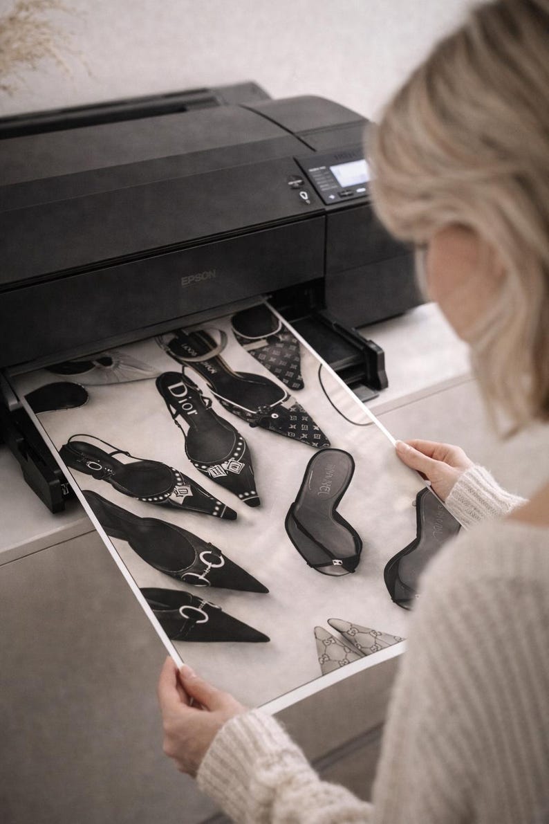 Puede incluir: Una impresora Epson negra imprimiendo una hoja de papel con ilustraciones de varios zapatos de tac&oacute;n negros y blancos. El papel presenta los logotipos de Dior y Chanel. La imagen es en blanco y negro.