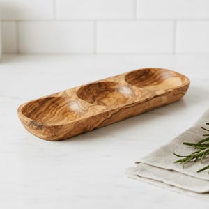 Peut inclure: Un plat de service en bois d'olivier avec trois compartiments ovales. Le plat présente un motif de grain naturel. Il est conçu pour servir des amuse-gueules ou des condiments. Une serviette en lin et des brins de romarin sont en arrière-plan.