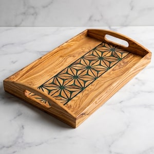 Peut inclure: Plateau de service rectangulaire en bois avec des poignées découpées. Le plateau présente un motif central incrusté de formes géométriques vert foncé. Le bois a un grain naturel et une couleur brun clair chaud. Environ 40 cm de long.