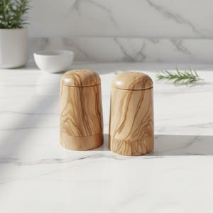 May include: Two wooden salt and pepper shakers with a natural wood grain pattern. The shakers are cylindrical with a rounded top and a small base. The tops have several small holes for dispensing the contents. The background is a white marble countertop.