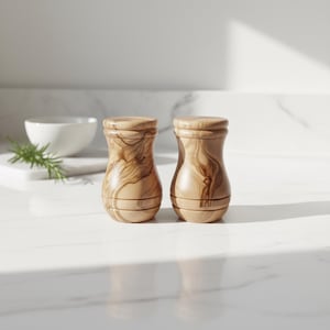 May include: Two wooden salt and pepper shakers with a light brown finish and unique wood grain patterns. The shakers have a classic hourglass shape and are displayed on a white countertop. A white bowl and sprig of rosemary are in the background.