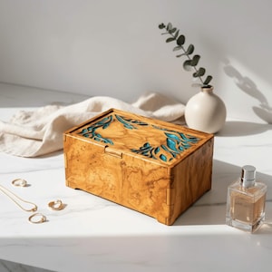 Pode incluir: Uma caixa de joias de madeira com acabamento em grão de madeira natural e detalhes turquesa. A caixa apresenta um design floral decorativo na tampa e tem formato retangular. Outros itens incluem joias, um frasco de perfume e um vaso pequeno.