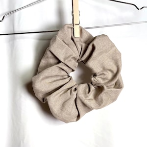 May include: A beige fabric hair scrunchie with a gathered, textured appearance, suspended from a silver metal clothes hanger by a small wooden clothespin. The scrunchie features a neutral, light brown tone and a soft, pleated circular design.