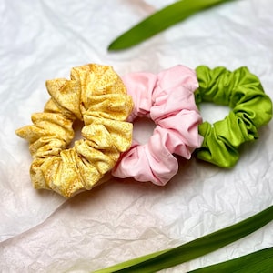 Puede incluir: Tres coleteros de tela, uno amarillo con estampado floral, uno rosa y uno verde, dispuestos sobre una superficie blanca texturizada. Los coleteros son circulares. Hojas verdes en el fondo.