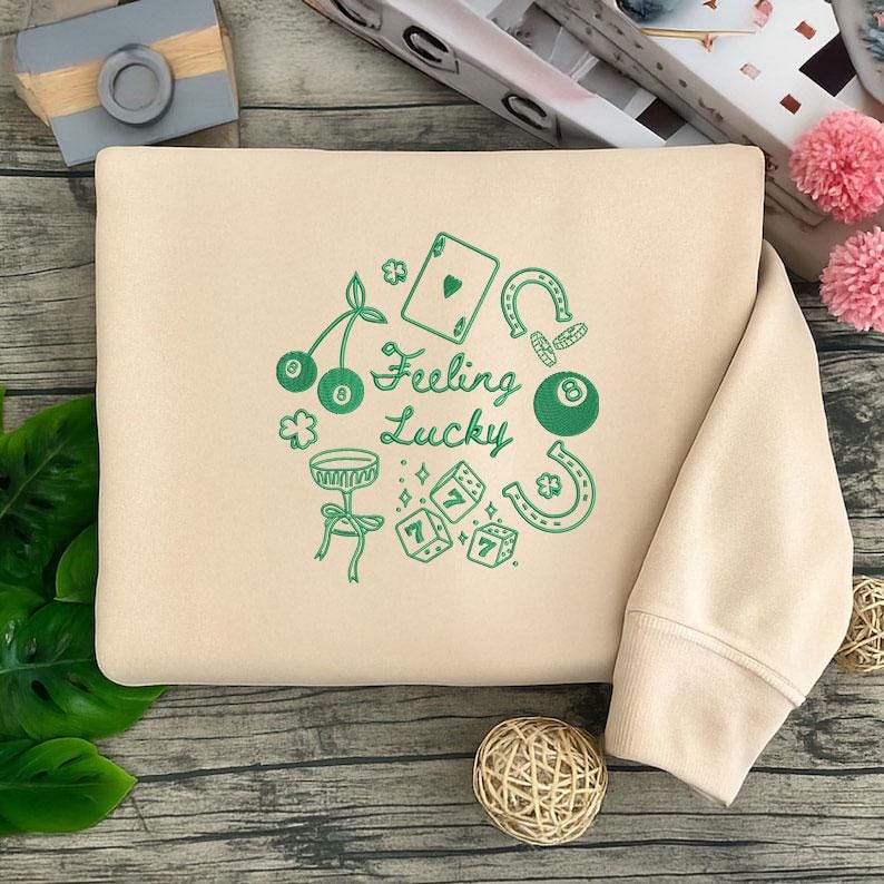 Pode incluir: Camisola bege com um design gr&aacute;fico verde. O design inclui cerejas, uma carta de jogar, uma ferradura, uma bola de bilhar, dados e as palavras "Feeling Lucky". A camisola tem gola redonda e mangas compridas.
