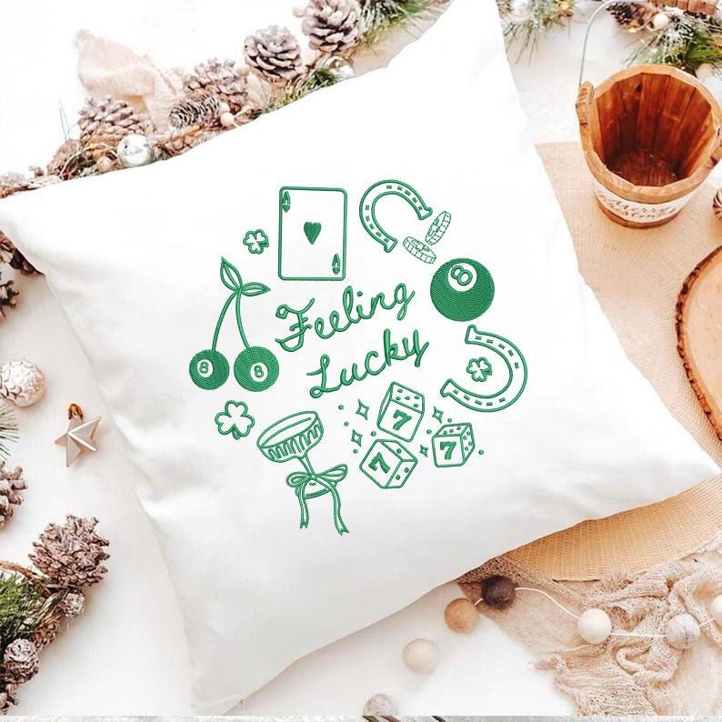 Pode incluir: Almofada quadrada branca com desenhos bordados em verde de cerejas, uma ferradura, dados, uma bola de bilhar e uma carta de jogar. As palavras "Feeling Lucky" est&atilde;o escritas em it&aacute;lico. Artigo decorativo.