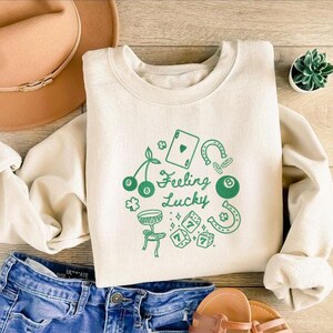 Pode incluir: Camisola creme com gr&aacute;ficos verdes e o texto "Feeling Lucky". O design inclui cerejas, uma carta de jogar, ferraduras, uma bola de bilhar, dados e um tambor. A camisola &eacute; combinada com cal&ccedil;&otilde;es de ganga azuis e um chap&eacute;u castanho.