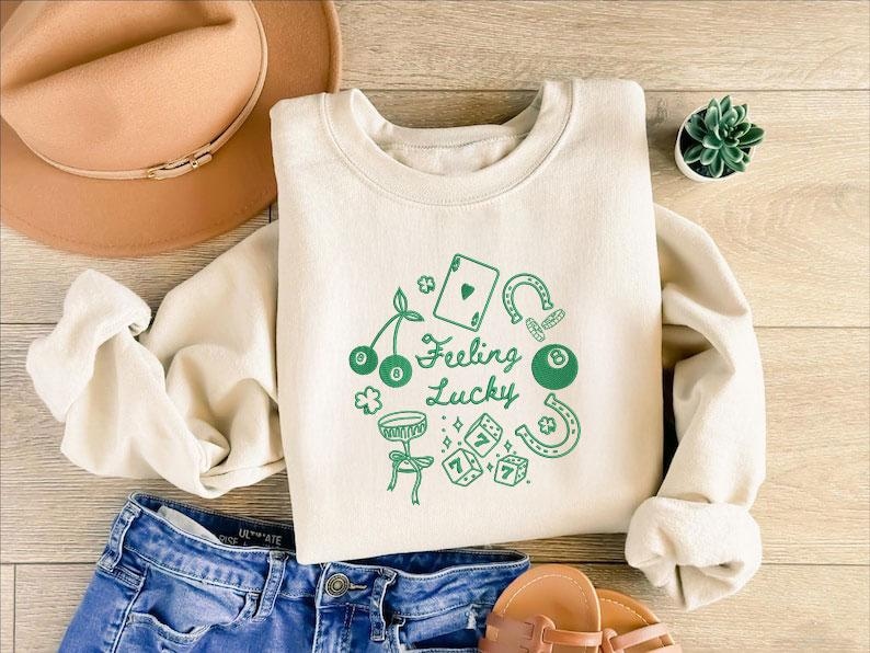 Pode incluir: Camisola creme com gr&aacute;ficos verdes e o texto "Feeling Lucky". O design inclui cerejas, uma carta de jogar, ferraduras, uma bola de bilhar, dados e um tambor. A camisola &eacute; combinada com cal&ccedil;&otilde;es de ganga azuis e um chap&eacute;u castanho.