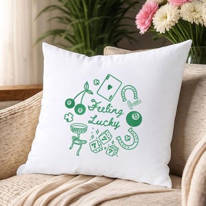 Pode incluir: Almofada quadrada branca com um gr&aacute;fico verde de &iacute;cones com tema de cassino, incluindo cerejas, uma carta de baralho, dados, uma ferradura e uma bola oito. As palavras "Feeling Lucky" s&atilde;o escritas em uma fonte de script. A almofada est&aacute; em uma superf&iacute;cie bege.