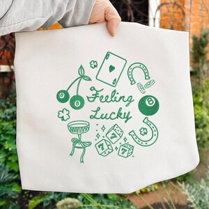 Pode incluir: Uma sacola branca com um design verde com o texto "Feeling Lucky" e v&aacute;rios s&iacute;mbolos da sorte, incluindo uma carta de baralho, dados, cerejas, uma ferradura e uma bola de bilhar.