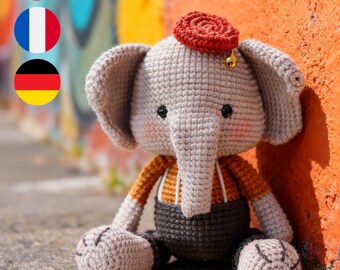 May include: A crocheted elephant doll in shades of gray, brown, and orange, wearing a red hat with a small gold bell. The elephant has black eyes and rosy cheeks. The doll is sitting against a colorful graffiti wall.