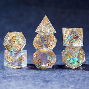 Può includere: Un set di dadi in resina trasparente iridescente per giochi da tavolo. I dadi hanno numeri color oro e un effetto olografico, con varie forme geometriche tra cui d4, d6, d8, d10, d12 e d20. I dadi sono disposti su una superficie blu.