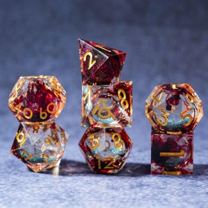 May include: A collection of polyhedral dice in various shapes, including a d4, d6, d8, d10, d12, and d20. The dice are clear with a red and gold interior and gold numbering. The dice are stacked on top of each other.