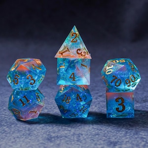Puede incluir: Un conjunto de siete dados poliédricos para juegos de mesa. Los dados son transparentes con una combinación de colores azul y naranja, y números dorados. Los dados están dispuestos sobre una superficie azul oscuro.