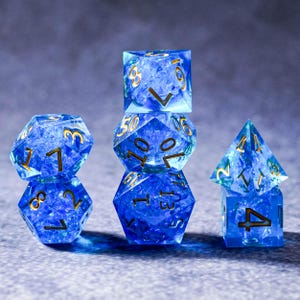 Puede incluir: Un conjunto de seis dados poliédricos azules con números dorados. Los dados son transparentes con un efecto veteado. Los dados están apilados, mostrando diferentes formas, incluyendo un cubo, un d10, un d6 y un d4.