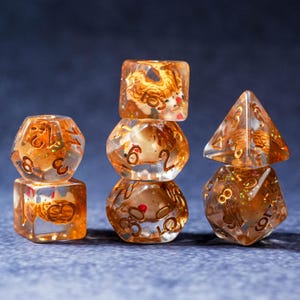 May include: Set of six clear resin dice with gold-colored numbers and embedded gold glitter and a tiger design. The dice are in various shapes, including a cube, a d8, a d10, and a d20. The dice are stacked on top of each other.