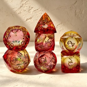 Può includere: Un set di sette dadi poliedrici traslucidi nelle tonalità del rosso, arancione e giallo. I dadi presentano numeri color oro e accenti glitterati interni. Sono disposti in tre pile, mostrando varie forme geometriche tra cui d20.