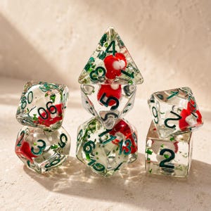 Può includere: Un set di dadi trasparenti in resina con dettagli rossi e verdi. I dadi presentano piccole forme rosse e fogliame verde, con numeri verdi. I dadi sono disposti in una formazione impilata.