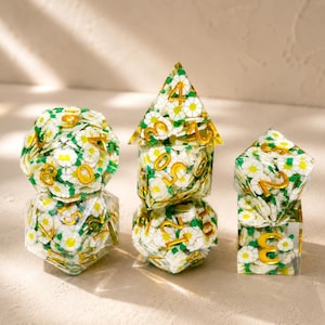 Può includere: Un set di sei dadi poliedrici per giochi da tavolo. I dadi sono trasparenti con un motivo floreale di margherite bianche e foglie verdi. I numeri sono dorati. Include un d4, d6, d8, d10, d12 e d20.
