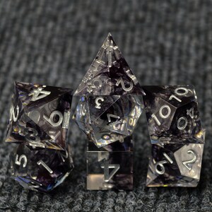 May include: A collection of clear, multi-sided dice with silver numbering. The dice have a dark, swirling pattern inside. The set includes a variety of shapes, including a pyramid, a cube, and other polyhedral forms, suitable for tabletop gaming.