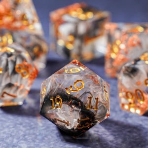 May include: Set of polyhedral dice with a clear resin base, swirled with black and white, and accented with gold numbering and orange glitter. The dice are designed for tabletop games.