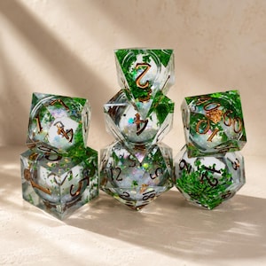 Può includere: Un set di sette dadi poliedrici per giochi da tavolo. I dadi sono trasparenti con fogliame verde e numerazione dorata. I dadi sono impilati, mostrando le loro diverse forme e dimensioni.