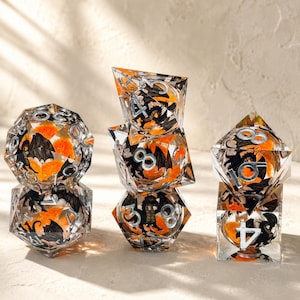 Può includere: Un set di dadi in resina trasparente con disegni di pipistrelli e fiori arancioni e neri. I dadi hanno numeri argentati e sono impilati, mostrando varie forme geometriche, tra cui un d20, d12, d8 e d6.