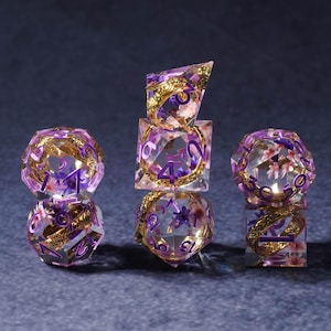 May include: A collection of clear resin dice with embedded purple flowers and gold accents. The dice are in various shapes, including a 20-sided die, a 6-sided die, and a 4-sided die. The numbers are in a purple font.