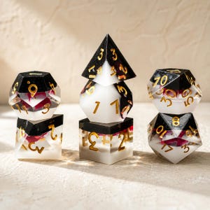 Può includere: Un set di dadi poliedrici per giochi da tavolo. I dadi sono realizzati in resina trasparente con dettagli neri e bianchi, numeri dorati e inclusioni floreali rosse. Il set include un d4, d6, d8, d10, d12 e d20.