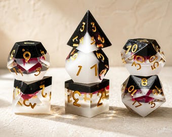May include: A set of polyhedral dice for tabletop games. The dice are made of clear resin with black and white accents, gold numbering, and red floral inclusions. The set includes a d4, d6, d8, d10, d12, and d20.