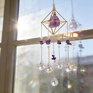 May include: A decorative suncatcher with a geometric gold frame holding a large purple amethyst crystal. Strands of clear beads, smaller amethyst pieces, and faceted crystal teardrops hang below, creating a shimmering effect. The suncatcher is hanging in front of a window.