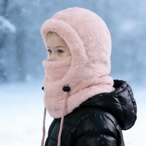 Può includere: Un morbido cappello invernale rosa con una protezione per il viso integrata. Il cappello ha una consistenza soffice ed è fissato con bottoni neri e cordini rosa. La persona nell'immagine indossa un cappotto invernale nero.