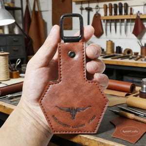May include: A brown leather keychain with a black metal loop and a hexagonal design. The keychain features a bull graphic and the text "HANDCRAFTED LEATHER GOODS". The item is held in a hand.