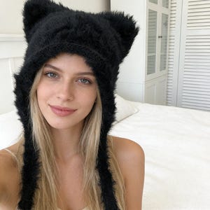 May include: A black, fluffy knitted hat with cat ears. The hat has long ties that hang down on either side. The hat is worn by a person with blonde hair and fair skin.