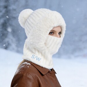 May include: A white knitted winter hat with a matching face covering. The hat has a fluffy trim around the face and a pom-pom on top. The face covering has the letters "S/W" embroidered in light blue.