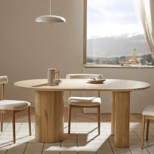 Pode incluir: Uma mesa de jantar oval de madeira clara com pernas cilíndricas e cadeiras a condizer. Uma lâmpada pendente branca pende sobre a mesa. As cadeiras têm assentos estofados de cor clara. Um tapete com um padrão de grelha está por baixo da mesa.