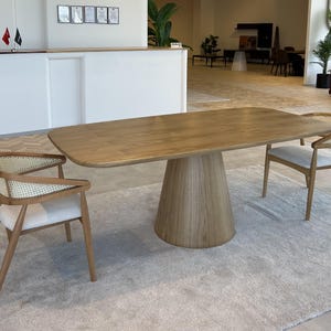 May include: A light brown wooden dining table with a rounded rectangular top and a conical base. Two wooden chairs with woven backs and light-coloured cushions are positioned on either side of the table. The table is set on a light-coloured rug.